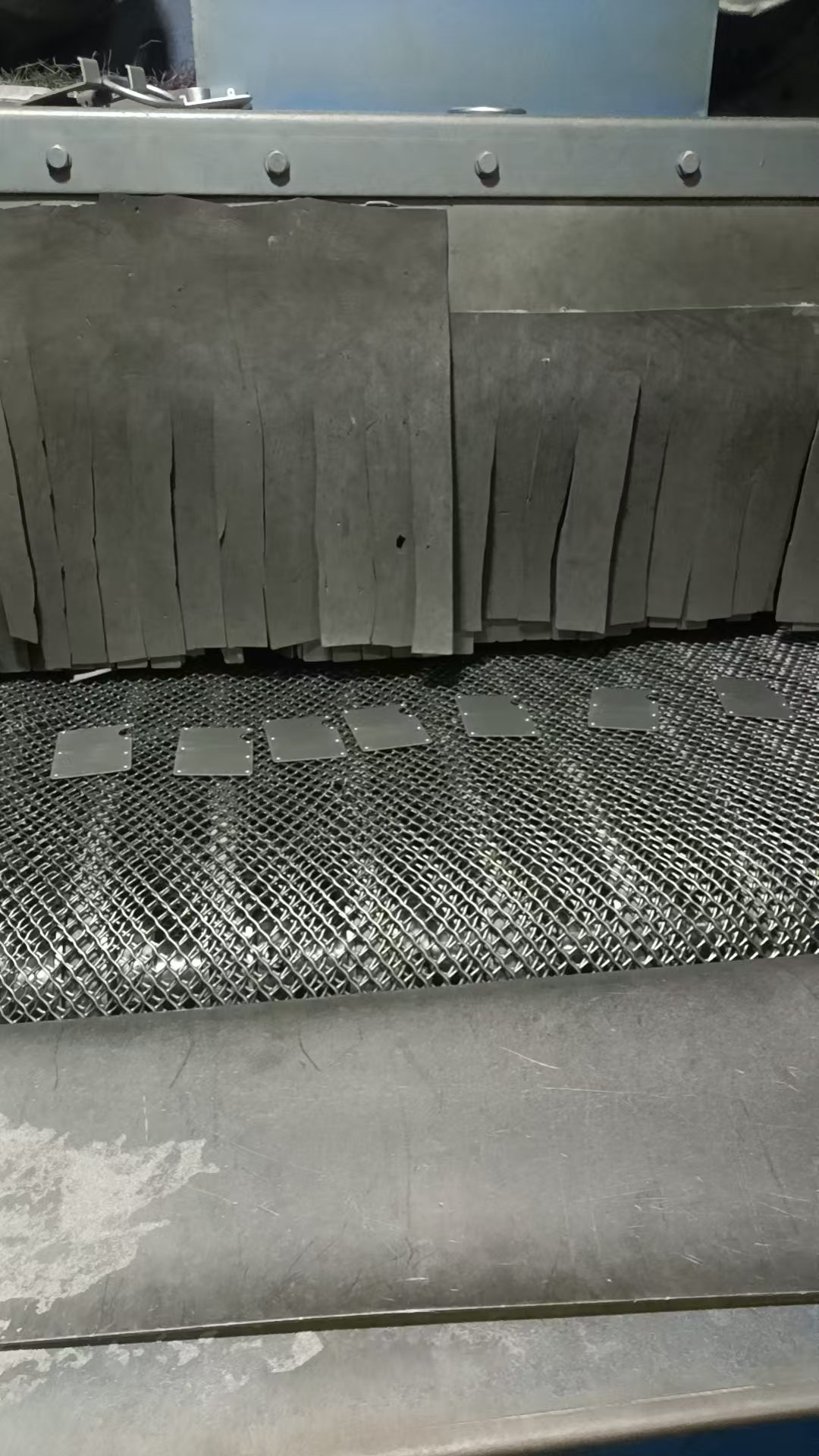 Stamped aluminium wallet shells feeding into a continuous mesh-belt shot-blasting machine