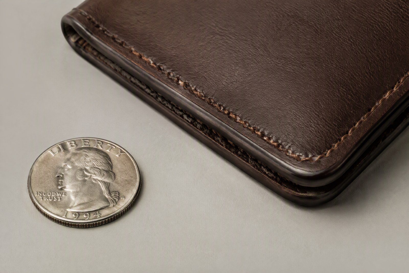 Close-up of a leather wallet edge showing burnished edge finishing and saddle-stitch details, a coin beside it for scale